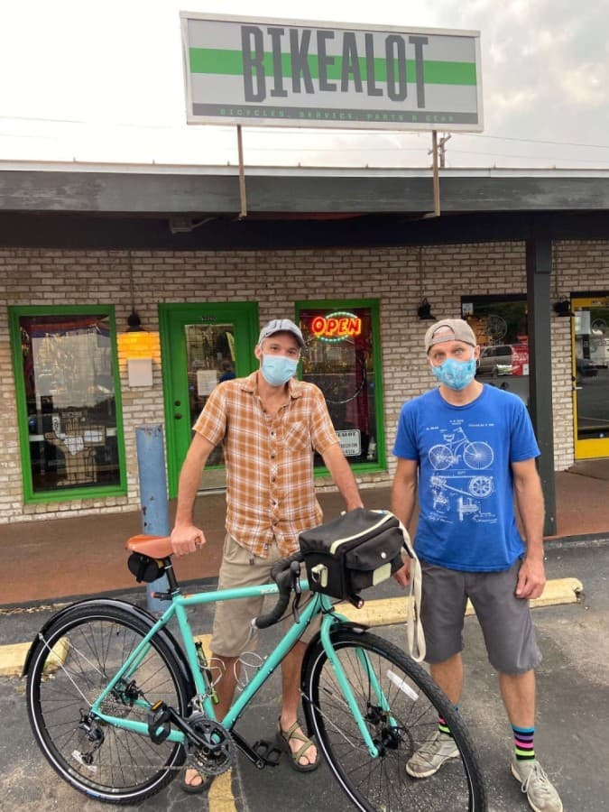 A photo of two men in masks standing with a new bike in front a shop with a sign that reads "Bikealot"