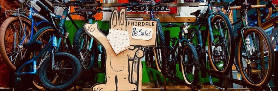 A photo of a carboard rabbit wearing a fabric mask and holding a sign that reads "be safe," posed infront of a rack of bikes