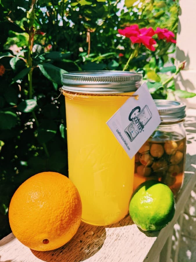 A photo of a bright yellow mixer in a mason jar next to fruit and olives