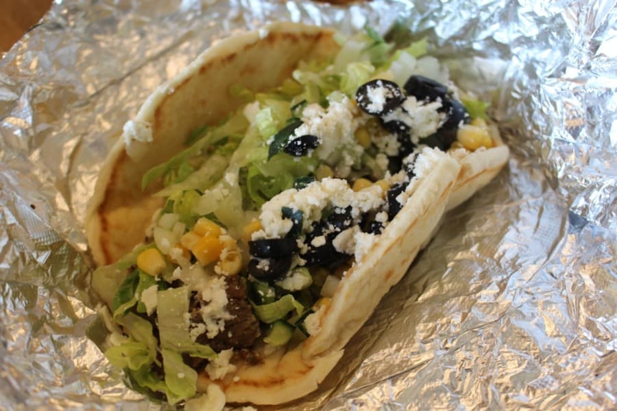 A gyro pita topped with lettuce, black olives, corn, cucumbers, onions, feta and creamy white sauce