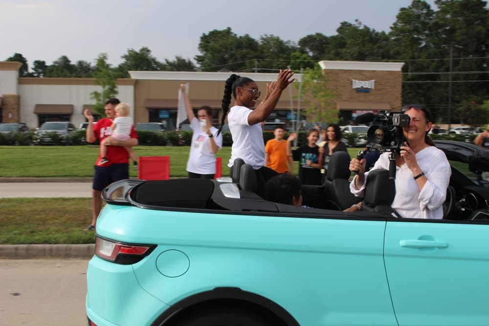 Biles smiles and waves from a convertible vehicle. (Ally Bolender/Community Impact Newspaper)