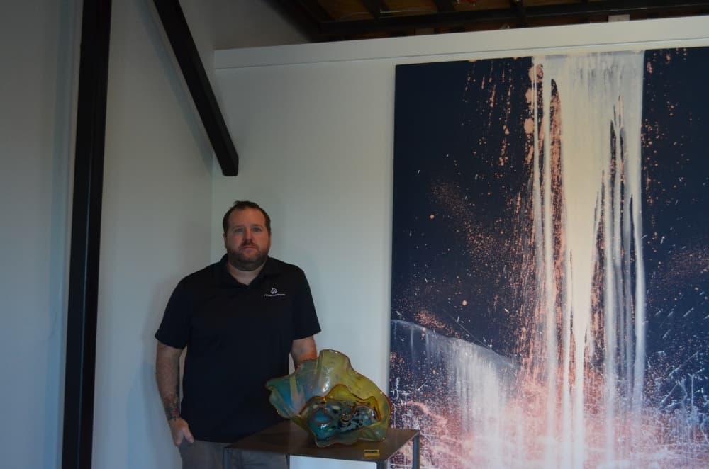 Owner Jeff Schneider poses next to one of his hand-blown glass pieces and a painting by artist Kevin Greer. (Amy Rae Dadamo/Community Impact Newspaper)