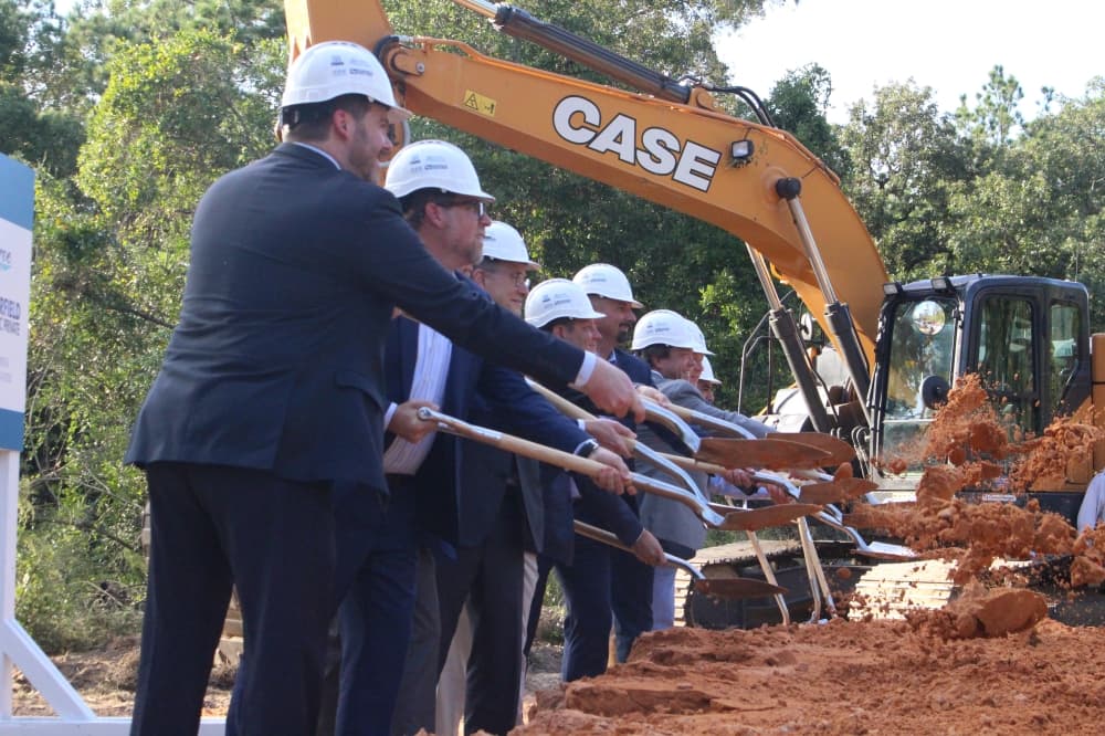 Men in suits and white hard hats toss soil with shovels