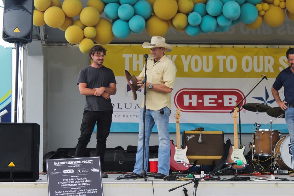 Mike Buchanan speaks on stage while holding a cowboy style hat next to artist Guido van Helten.