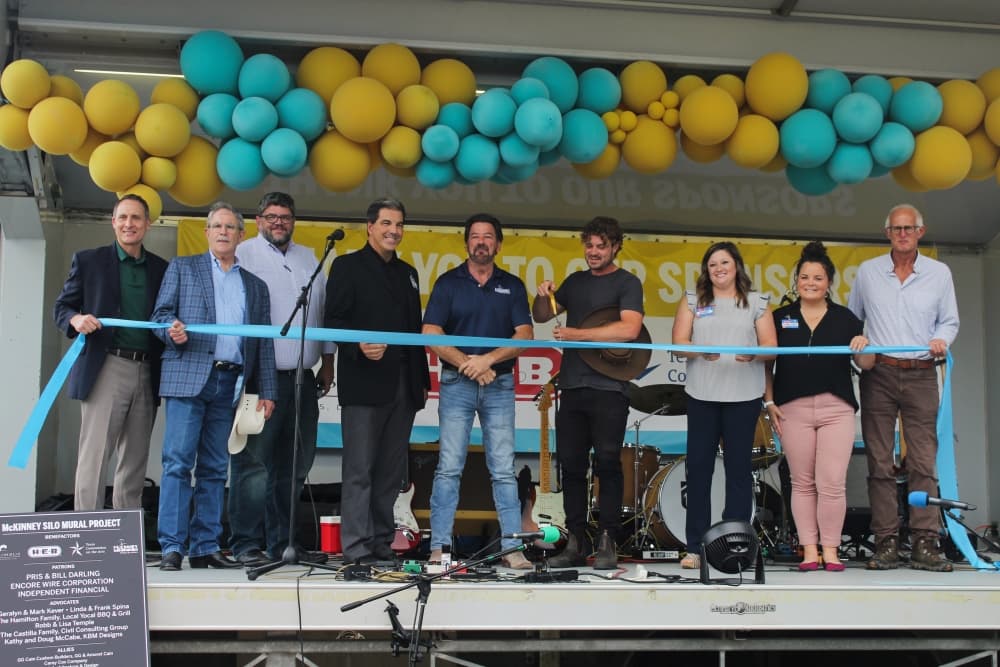 A group of people on a stage hold a ribbon while the honored artist cuts it.