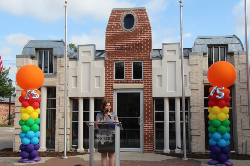 A woman speaking at a podium in front of the Safety Town replica of Frisco City Hall.
