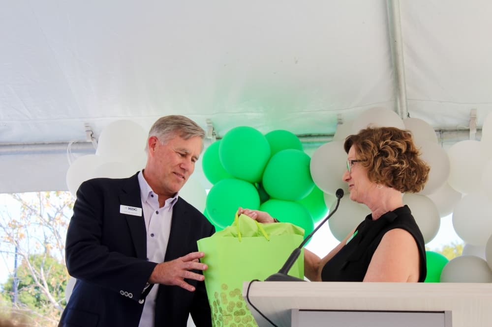 Lynne McLean giving a green gift bag to Bill Guthrey at a podium in front of green and white balloons.
