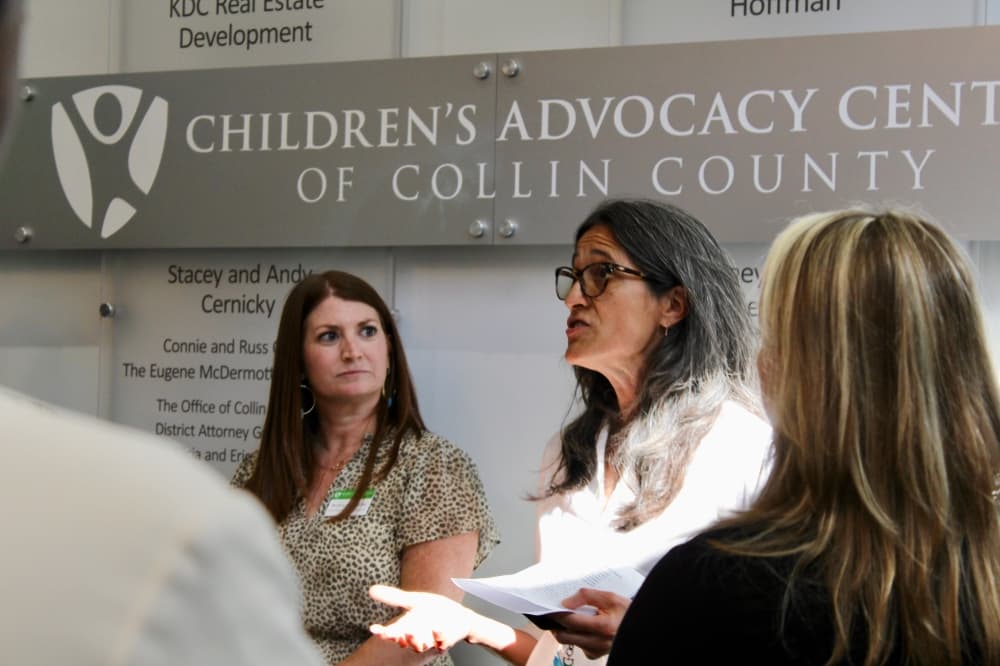 A woman speaking to a group in front of a sign with the organization's name.