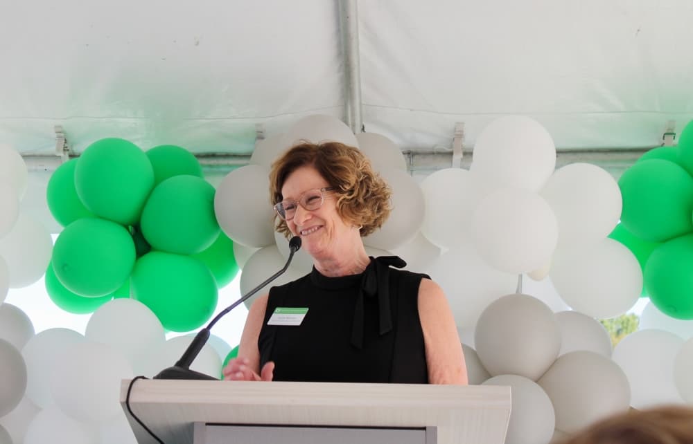 Lynne McLean at a podium in front of green and white balloons.