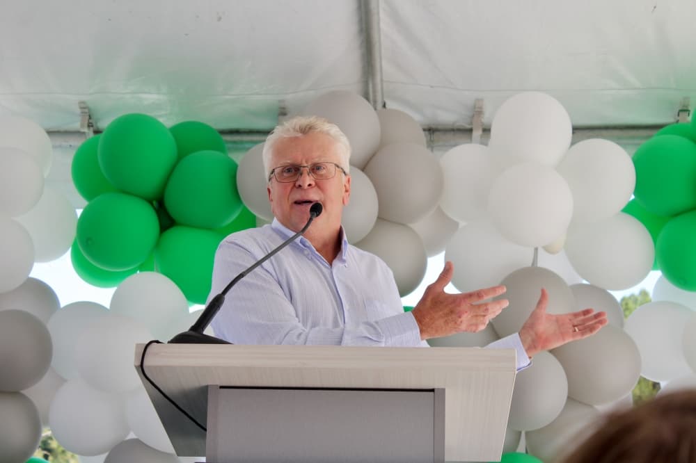 Mark Speese at a podium in front of green and white balloons gesturing to the building.