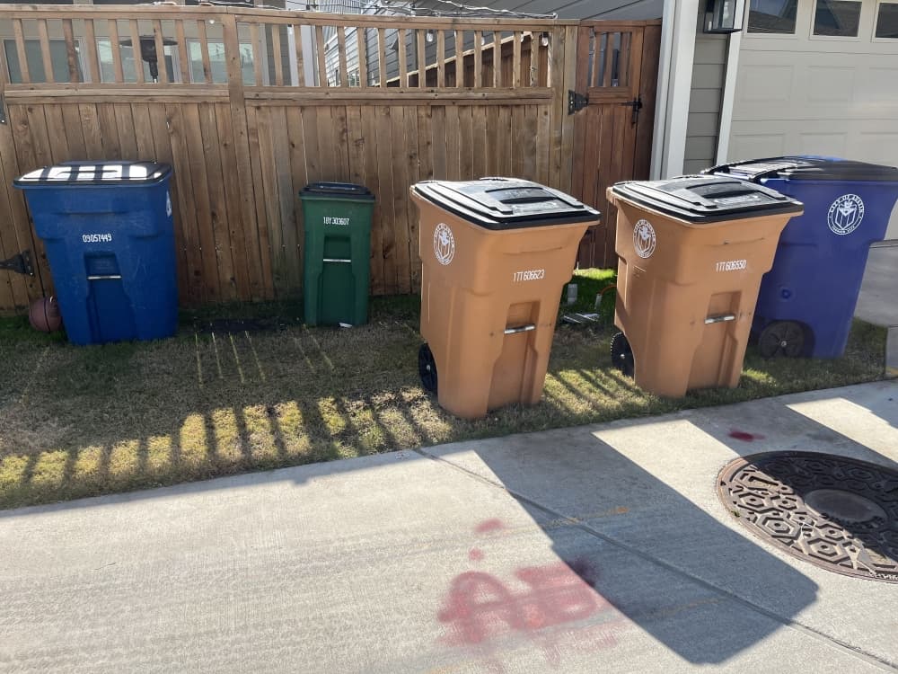 composting, recycling and trash bins sit in a grass patch in between a drivewaay and fence