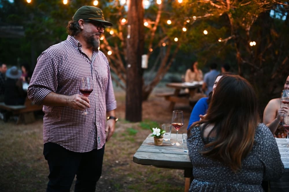 man in hat stands next to table holding a glass of red wine 