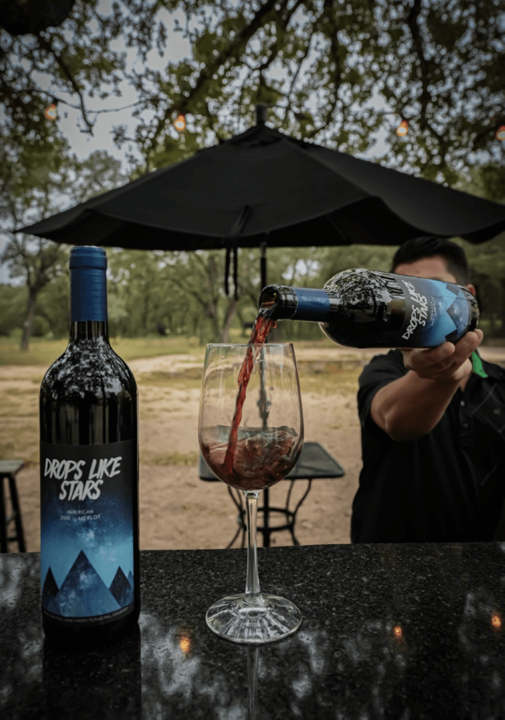 man pours wine into a wine glass with umbrella and trees in the background