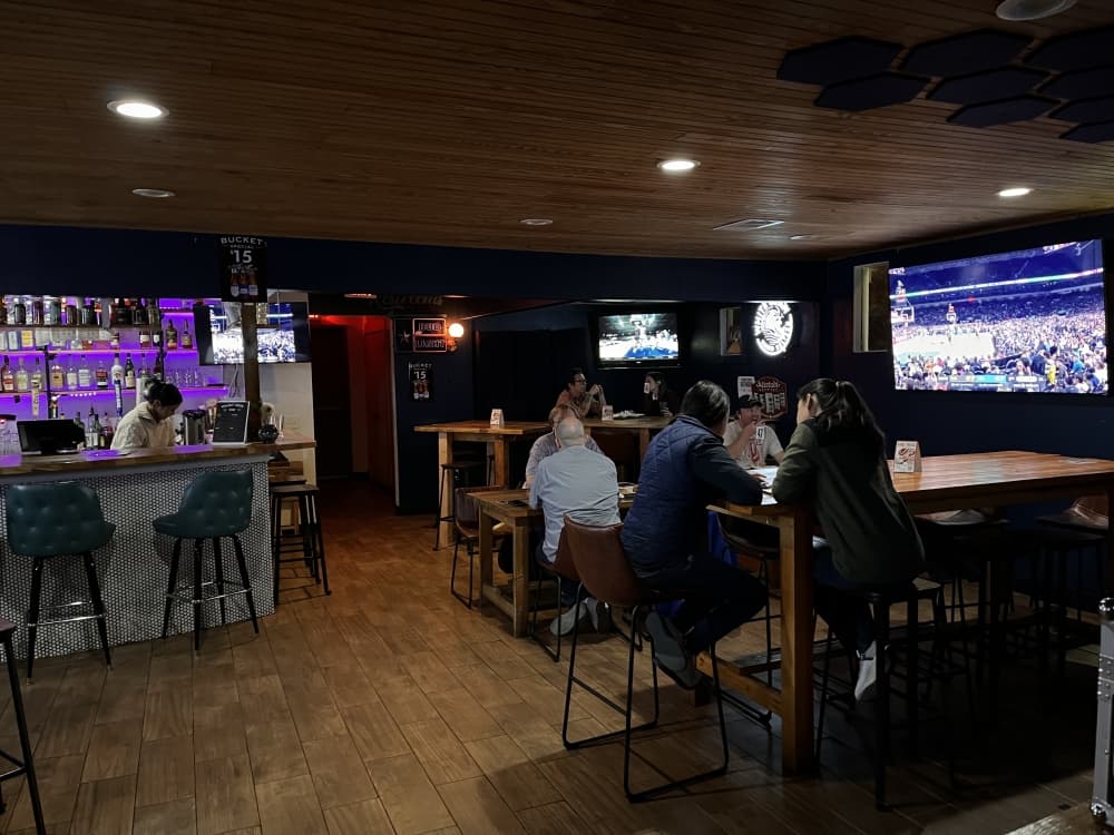 Bar and a television screen with customers sitting on chairs and a table.