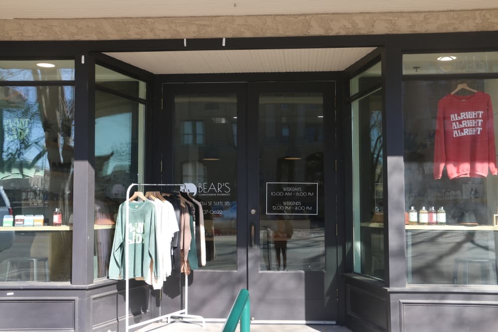 the storefront has glass windows with black trim and clothing on a rack beside the front doors 