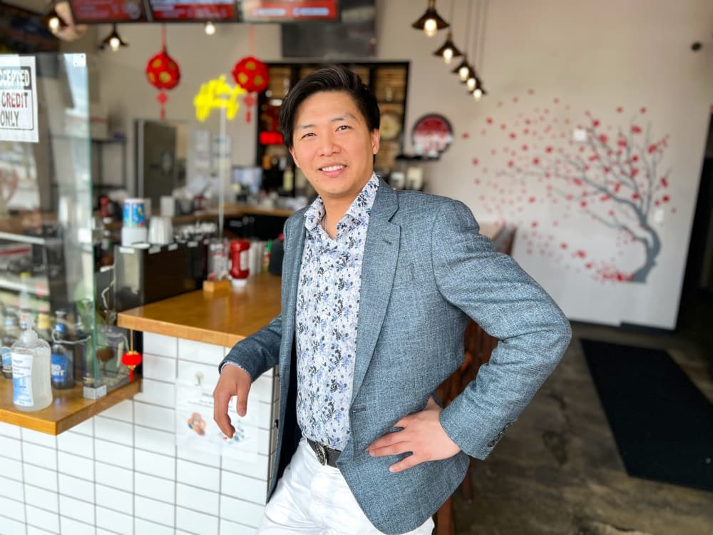 owner stands in grey jacket and printed shirt with hand on hip 