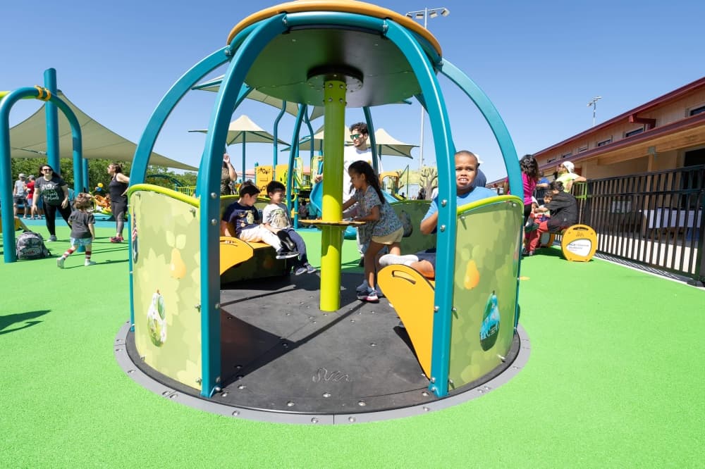 a group of children in a covered area on a playground with other children and adults in the background also engaging with the playground space