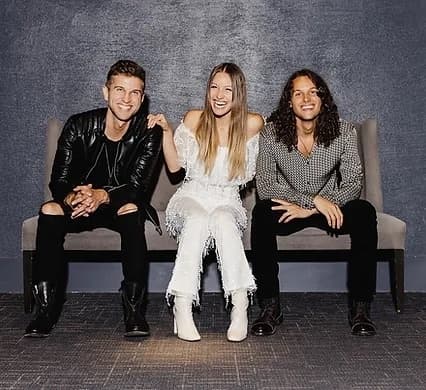 Three people sit on a bench in front of a grey wall; the woman in the middle is wearing white.
