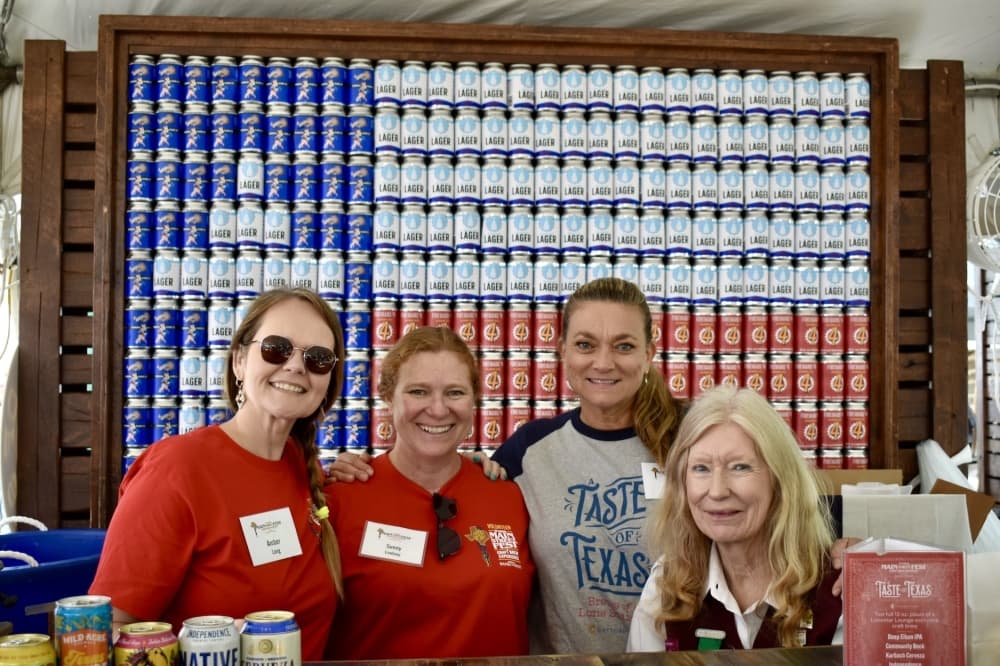 four women smile in front of a Texas flag made of beer cans