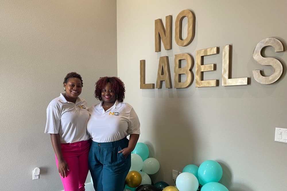 Founders Vallerie Dean and Erica Jackson standing in front of balloons and a "no labels" wall plaque.