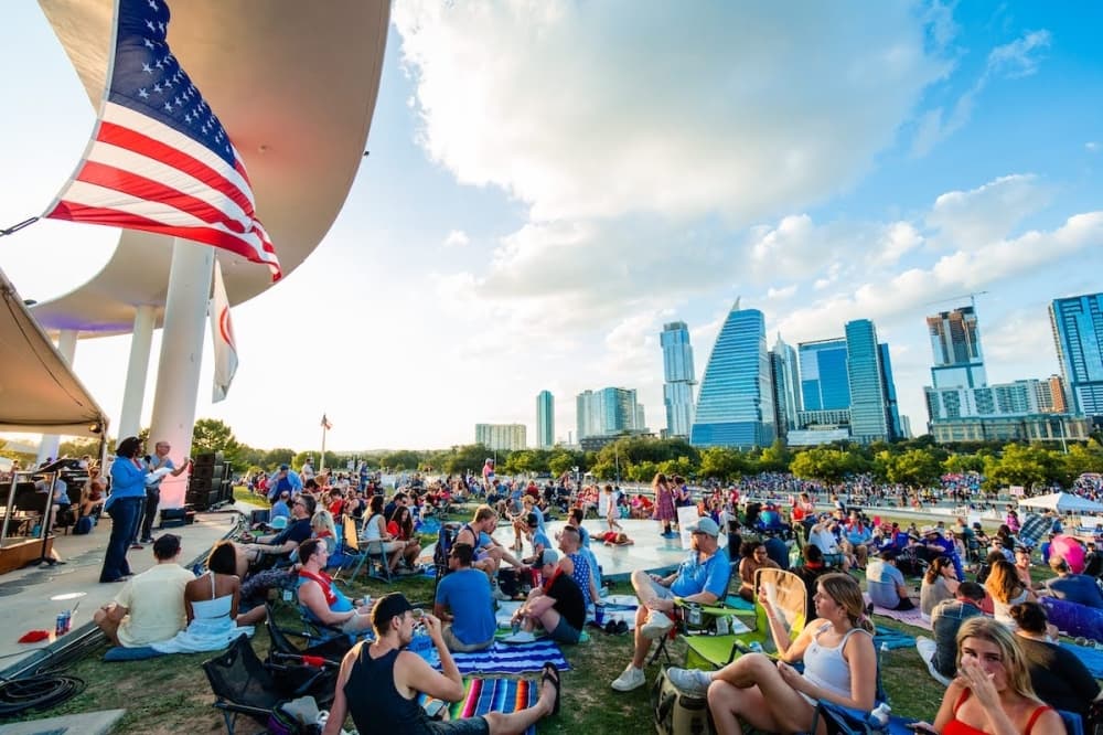 A crowd gathers by a flag with the Austin skyline in the background on a sunny day