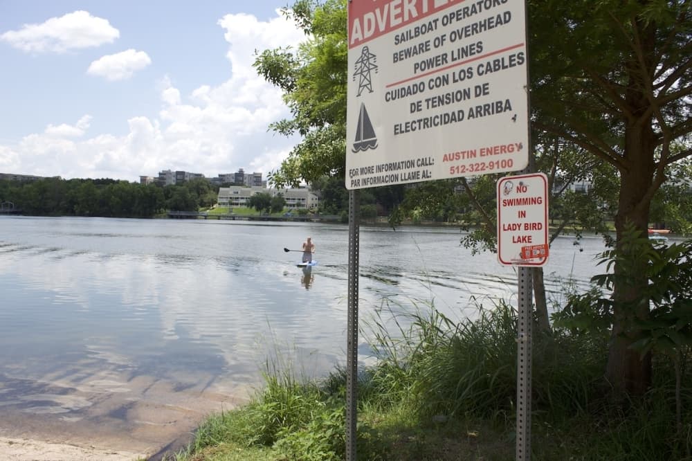 sign reads no swimming in lady bird lake with someone paddleboarding in the background