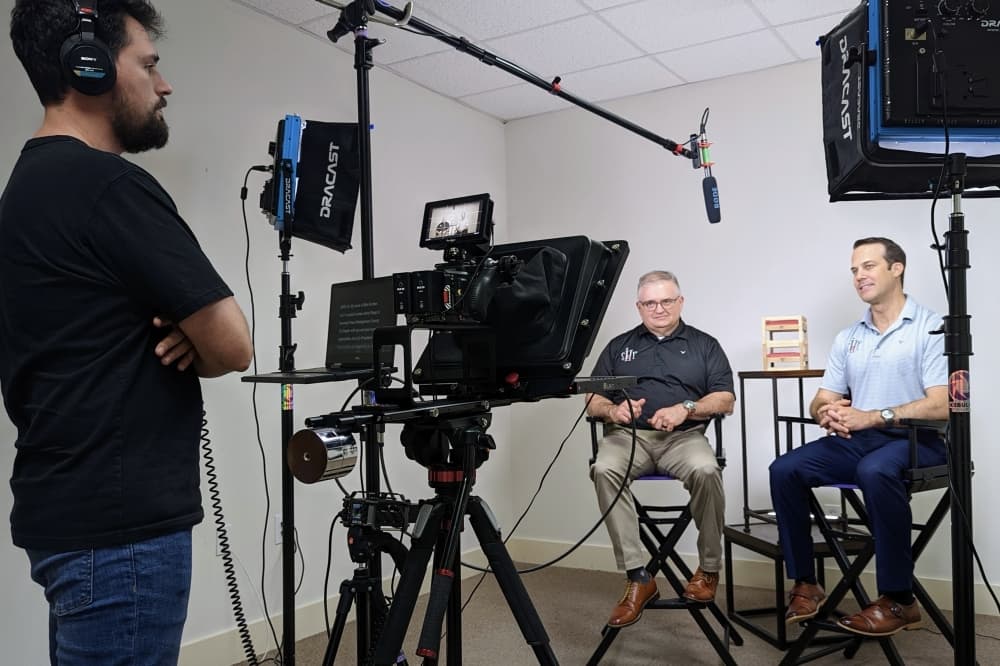 two people sit for an interview while one person stands behind a camera