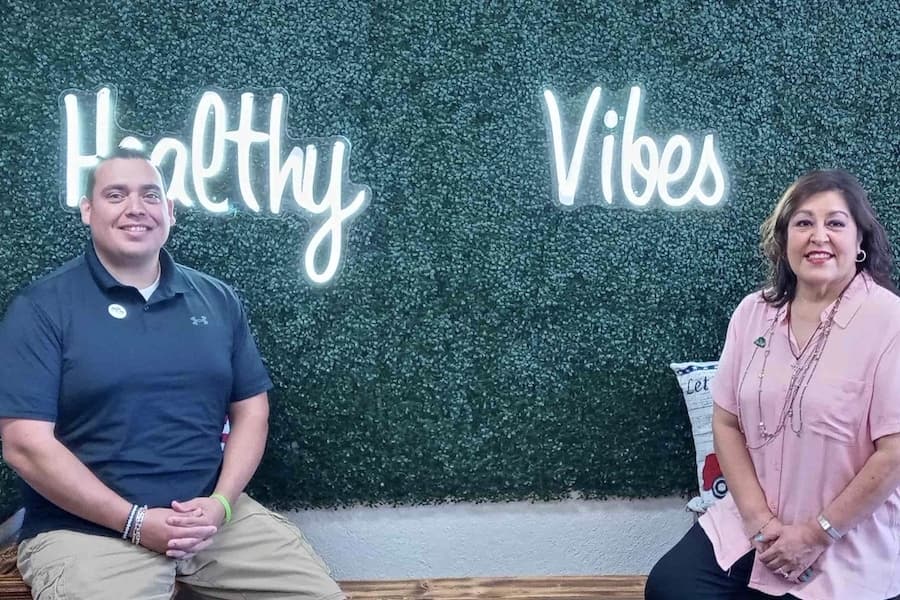 Anthony Bernal and Delma Ferris sit in front of a sign that says healthy vibes