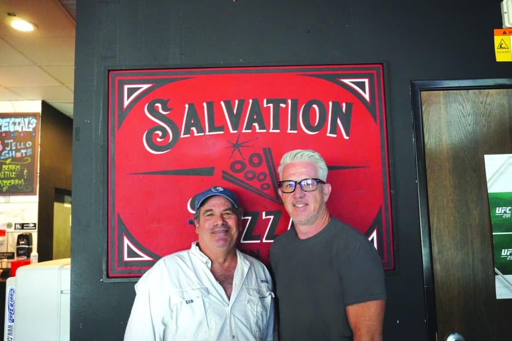 2 men standing in front of "salvation pizza" sign 