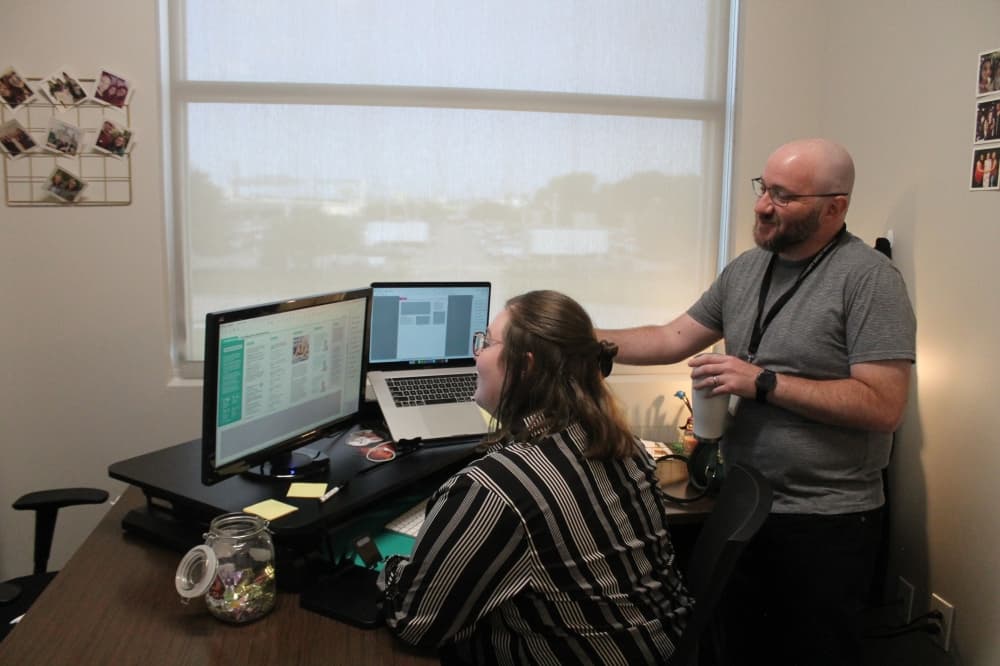 kaitlin schmidt sits at a desk with two monitors and matt stephens stands behind her pointing at one of the screens