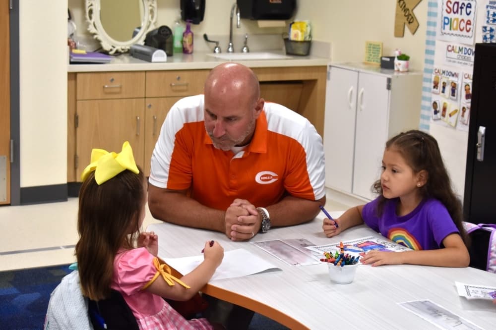 a man kneels by a table and speaks with two young girls