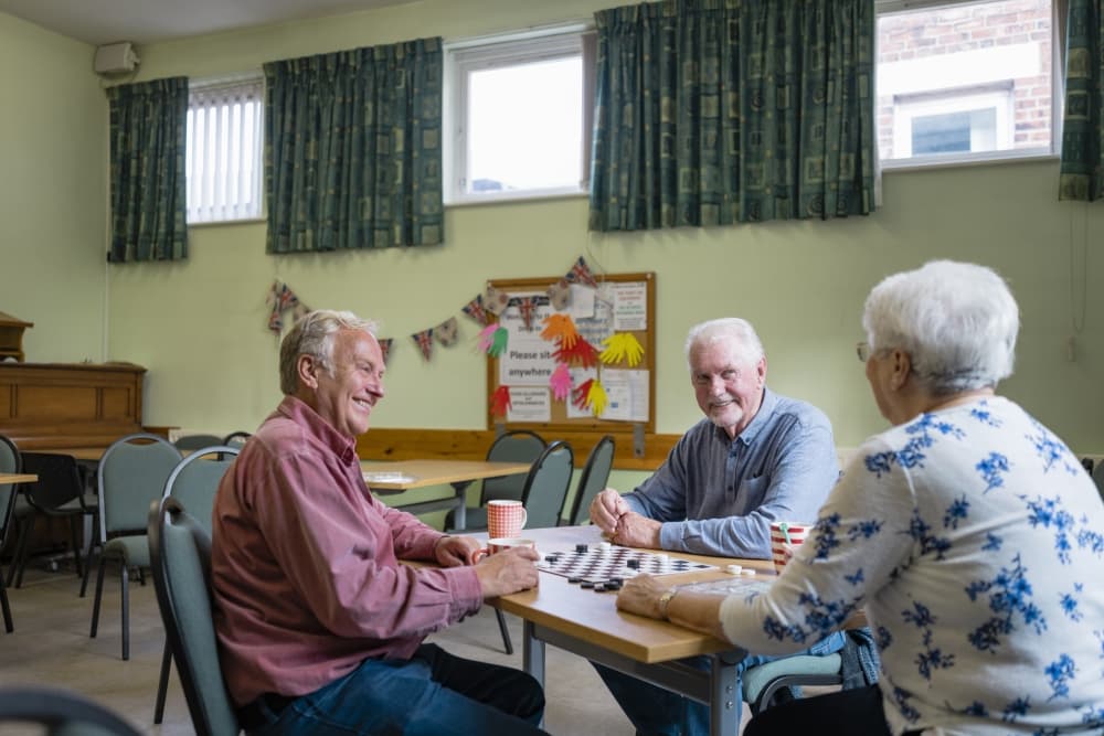 A shot of a small group of people visiting a warm hub/food bank which is a safe place for people to enjoy a warm and friendly environment in the community with the current cost of living increasing. Three senior friends are sitting around a table, they are playing a game of checkers.