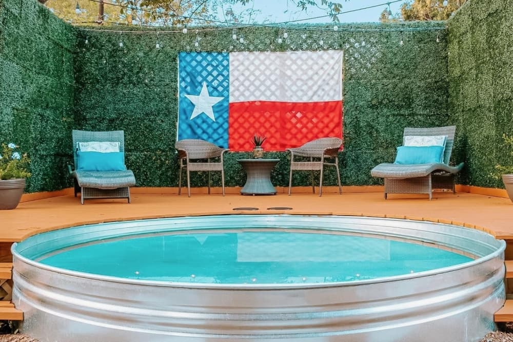 A stock tank pool with a wooden deck built around it and some patio chairs nearby.