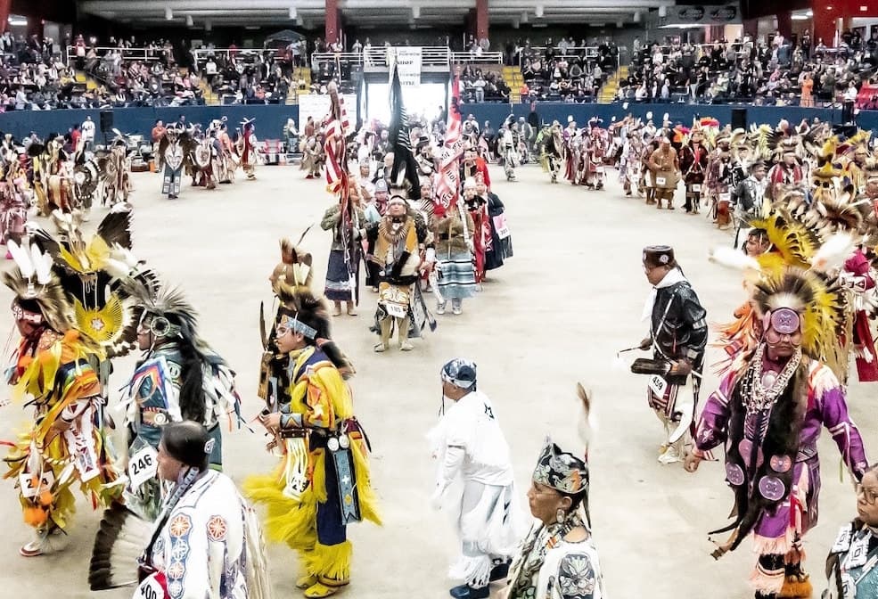 A group of people in traditional Native American attire dance around the center together inside.