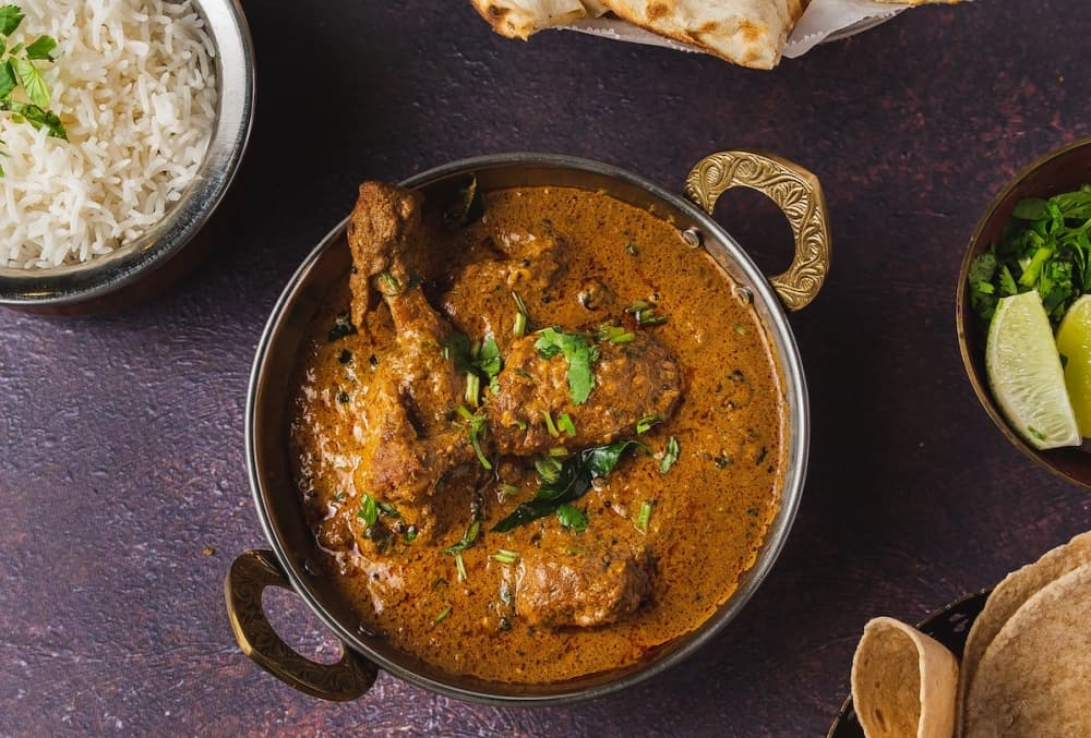 A bowl of chicken curry from Sangam sits on a table with sides of rice and bread beside it.