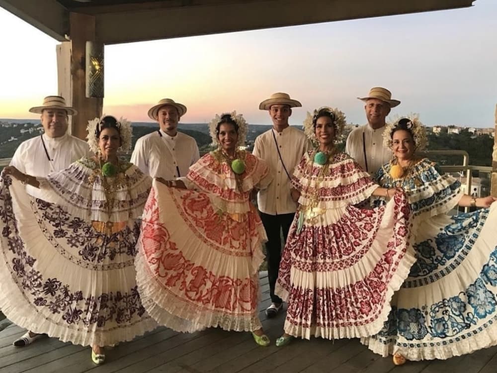 Four women dressed up in colorful, long dresses in between four men wearing white shirts and hats