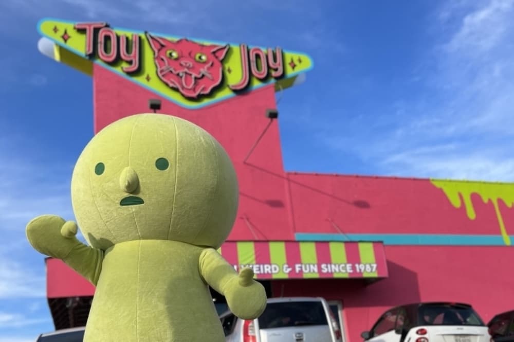 A giant green mascot waves to those passing by outside of the new Toy Joy location on a sunny day.