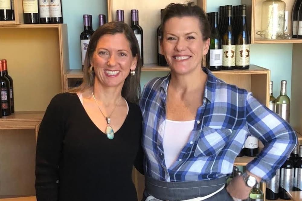 The owners stand next to each posing in front of a wall of wine.
