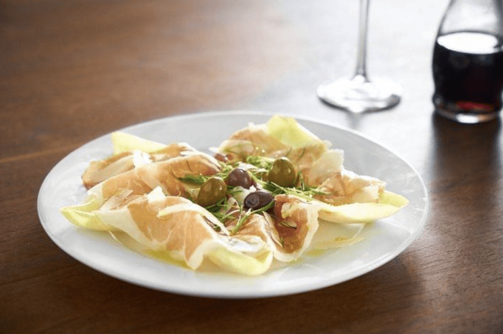 A plate of prosciutto with shaved Parmesan Reggiano, truffle oil, olives and green onion sits on a table.