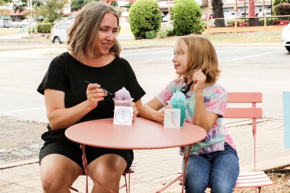 A child and woman sit at a dining table outside of the food truck and eat snocones.