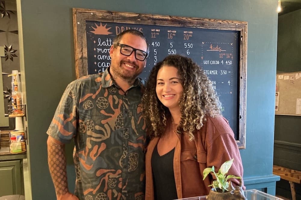 Matt and Brandy Stone pose for photo in front of coffee menu on a a chalkboard. 