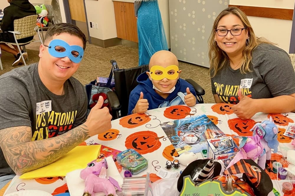 at a table, two adults and a child give thumbs up. An adult and the child are wearing superhero masks over their eyes