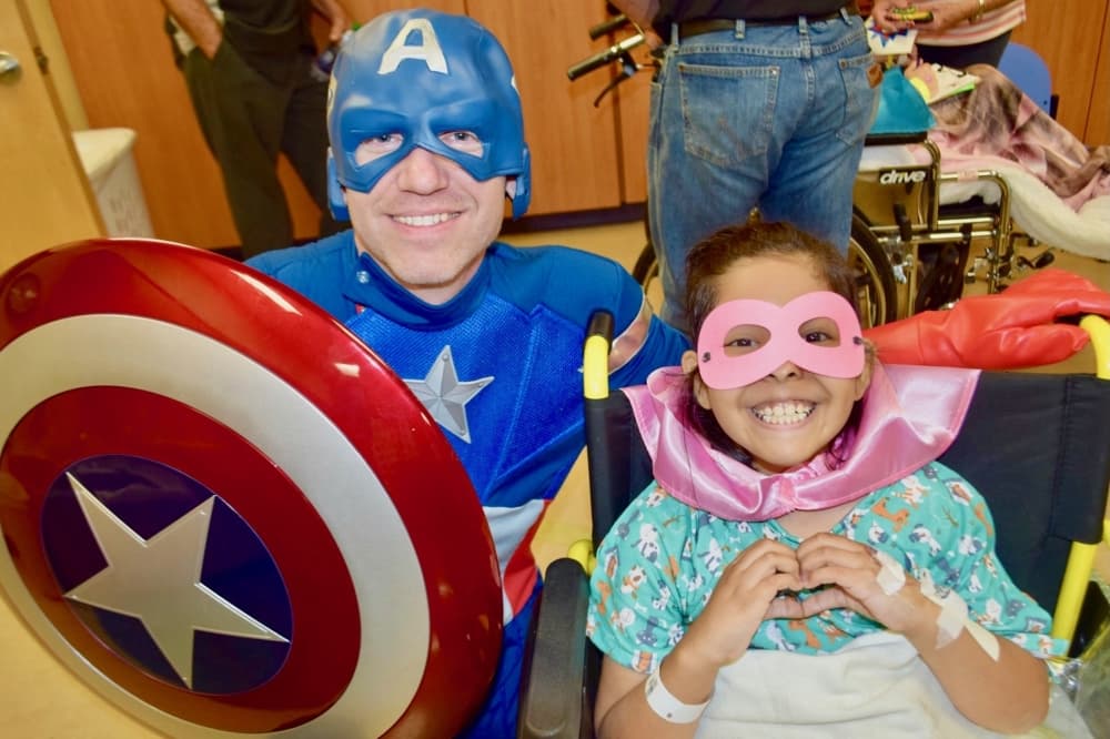 a child in a wheelchair wearing a superhero mask over her eyes poses for a photo next to someone dressed as Captain America