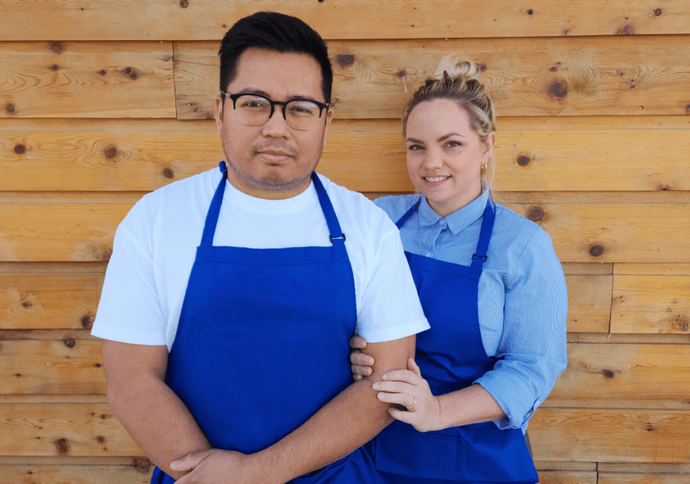 The owners wear blue aprons and stand in front of a wooden wall.