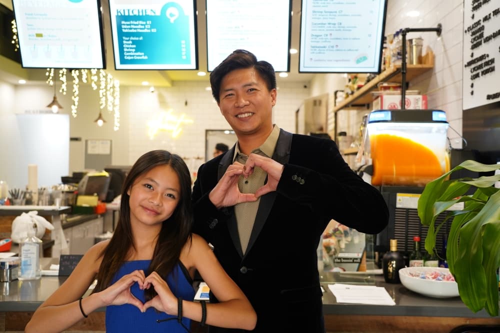 Ethan Huynh stands behind his daughter, Ava, in the restaurant while both make heart signs with their hands.