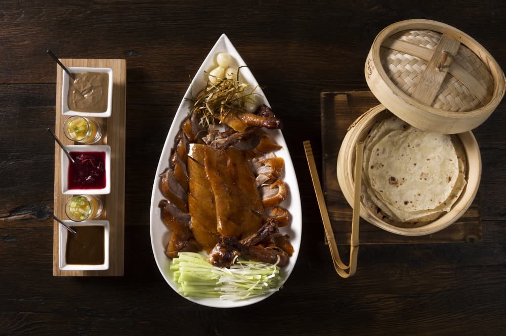 A dark table holes a white plate with sliced peking duck, and to either side is a tray with three sauces and a container with homemade pancakes.
