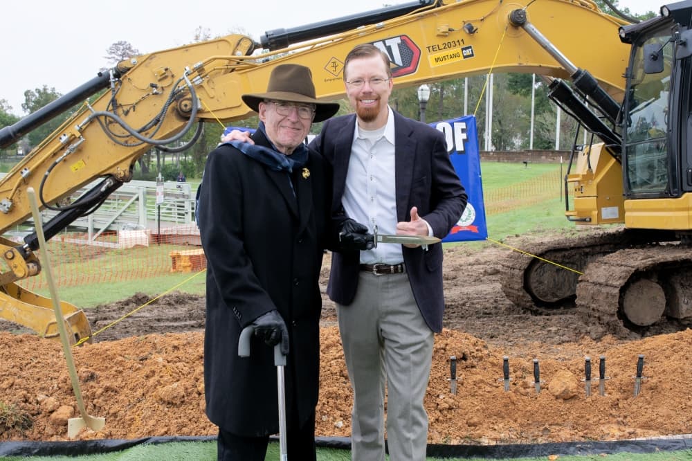 Two men stand in front of a piece of construction equipment 