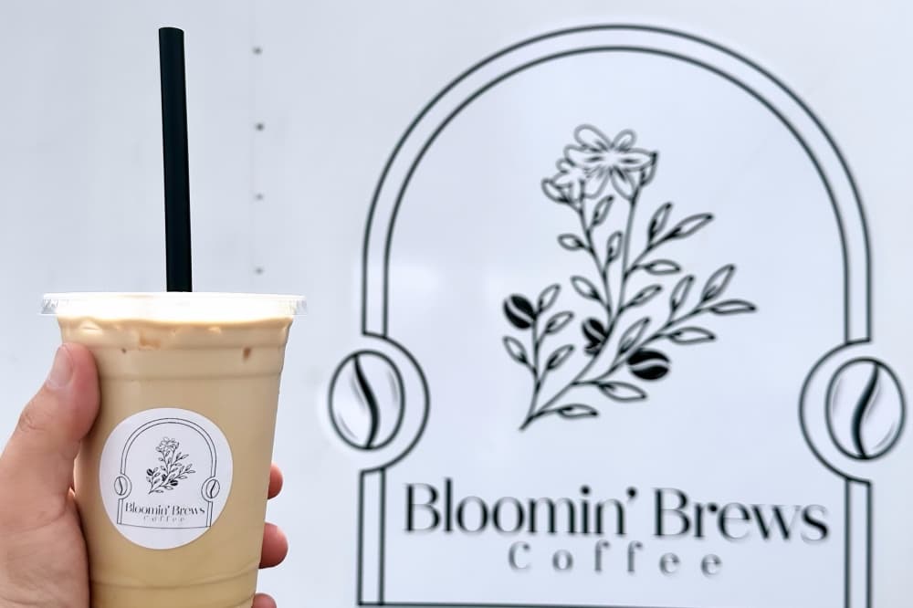 an iced coffee held up in front of the Bloomin' Brews sign