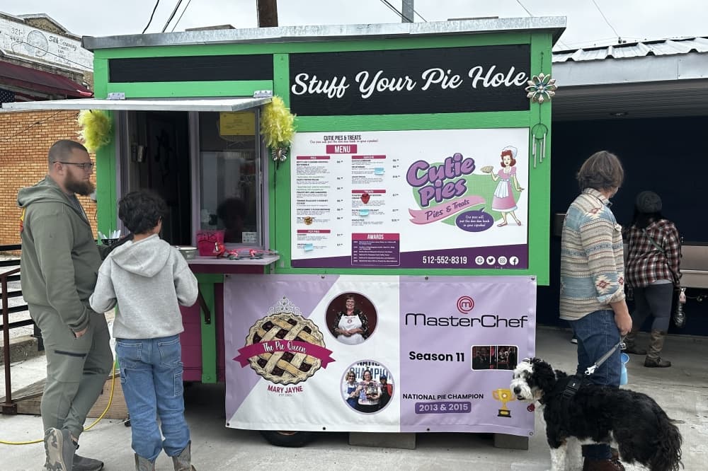 The green food trailer is surrounded by people and a dog.