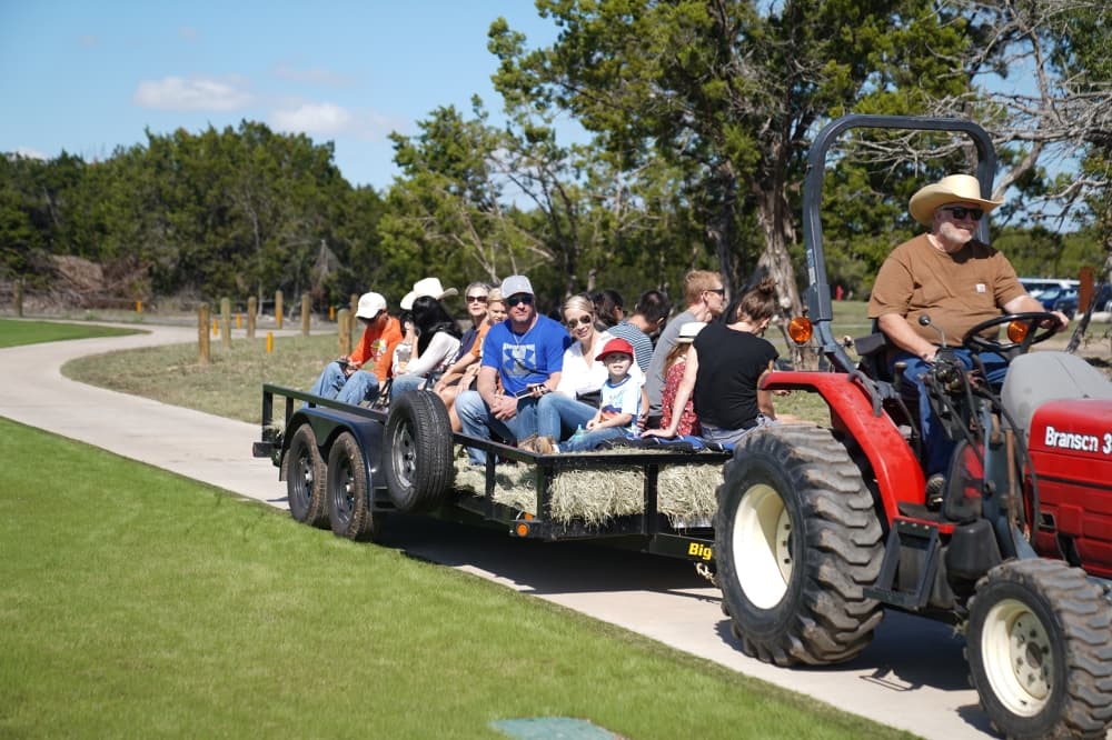 A group of people are loaded on trailer with hay while being pulled by a person in a cowboy hat on a red tractor.